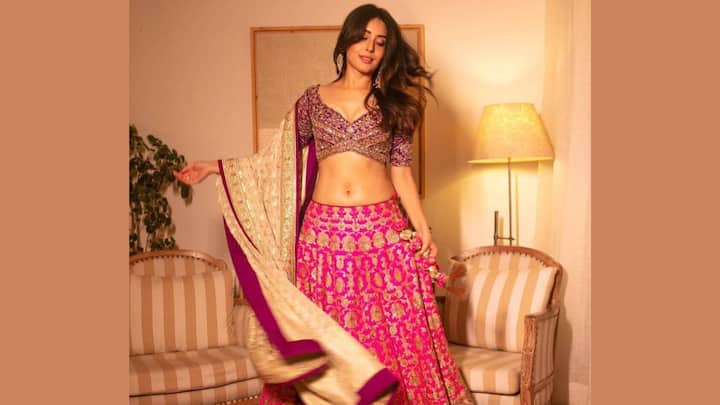 Pink Lehenga With Purple Blouse: Kritika Kamra is pictured wearing a vibrant festive Indian ensemble, specifically a lehenga choli. The outfit features a deep pink or fuchsia flared skirt (lehenga) with intricate gold embroidery and a matching, short-sleeved, deep-cut blouse (choli) with heavy gold and sequin embellishment. Pairing the lehenga with a cream- or beige-coloured dupatta, which has a thick magenta and gold border, she looks glamorous. (Image Source: Special Arrangement)