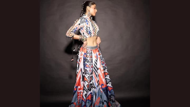 Gipsy Lehenga: Kritika Kamra is seen wearing a vibrant, contemporary take on the traditional Indian lehenga choli. The outfit features a high-neck, full-sleeved crop top and a floor-length A-line skirt or lehenga. Both pieces are adorned with a striking, multi-colour, abstract, or possibly Ikat-inspired ethnic print in shades of orange, red, blue, black, and white. The top is a form-fitting choli that exposes the midriff, and the sleeves have cutout or lattice details with tassel accents. Her look is accessorised with heavy, oxidised silver jewellery, including large jhumka-style earrings and a stack of bangles on both wrists, some of which also have tassels. (Image Source: Special Arrangement)