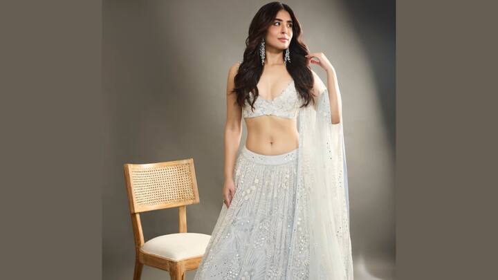 Pastel Blue Lehenga With Thread Work: Kritika Kamra embodies ethereal elegance in a soft pastel blue lehenga choli, perfect for a subtle yet shimmering festive look. The ensemble features a lightly hued skirt and a deep-neck, barely-there choli, both heavily adorned with intricate white embroidery, floral motifs, and scattered mirror work, catching the light beautifully. She completes the dreamy attire with a sheer dupatta draped over one arm and accessorises with dramatic silver or diamond chandelier earrings. (Image Source: Special Arrangement)