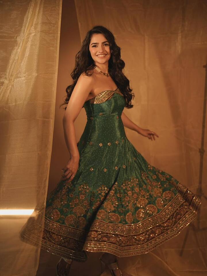 रुक्मिणी ने एक गहरे हरे रंग का खूबसूरत ट्रेडिशनल गाउन पहना है. यह गाउन स्ट्रैपलेस है और इस पर गोल्डन धागों और सीक्वेंस का बहुत बारीक काम किया गया है. गाउन का स्कर्ट काफी घेरदार है और उस पर भी बॉर्डर पर भारी कढ़ाई है. उन्होंने गले में एक पतला-सा गोल्डन चोकर नेकलेस और हल्के कर्ल वाले खुले बाल रखे हैं.