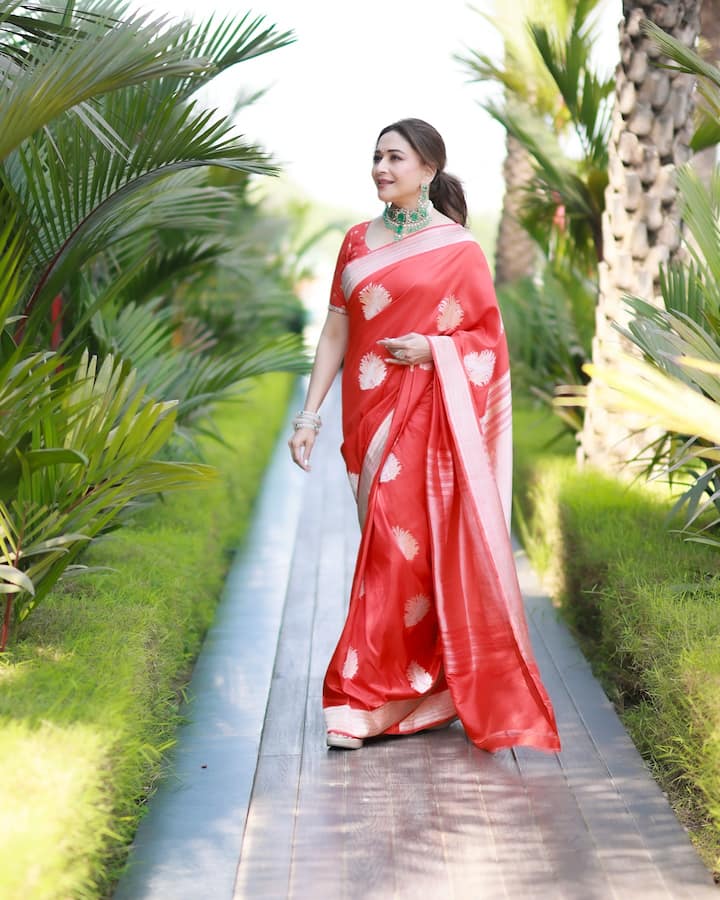 Madhuri is looking absolutely royal in this red orange colored silk saree. The light golden colored Banarasi booti work in the saree is giving it a classic and festive touch. She has paired this saree with a contrast jewelery green colored choker necklace and matching earrings, which is adding a new color to the entire look.