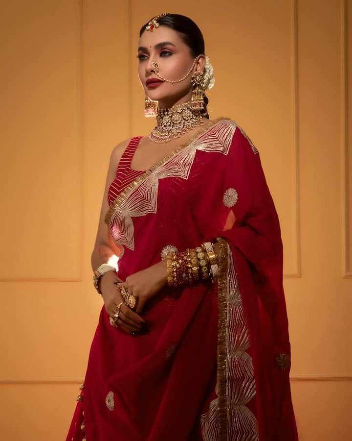 पवित्रा पूनिया का यह लुक एक शानदार ट्रेडिशनल ब्राइडल अवतार है. उन्होंने लाल रंग का लहंगा पहना है, जिस पर ज़री और गोटा पट्टी का काम किया गया है. लहंगे के साथ मैचिंग वेलवेट का दुपट्टा है, जिसकी बॉर्डर पर हैवी एम्ब्रॉइडरी है. इस लुक की जान उनकी हैवी ट्रेडिशनल ज्वेलरी है: एक चोकर नेकलेस, बड़े झुमके, माथा पट्टी, नोज रिंग, और दोनों हाथों में कई कड़े. उन्होंने अपने बालों का स्लीक बन बनाया है और डार्क लिपस्टिक के साथ ड्रामाटिक मेकअप रखा है.