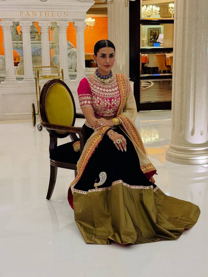 पवित्रा का यह लुक एक रॉयल एथनिक अंदाज़ है. उन्होंने तीन रंगों का भारी लहंगा पहना है. ऊपर रानी पिंक रंग की ब्लाउज है जिस पर सफेद और सघन कढ़ाई की गई है. लहंगा काला वेलवेट का है जिसके नीचे गोल्डन-ऑलिव रंग का बॉर्डर है. उन्होंने अपने इस आउटफिट को बेज और गोल्डन रंग के दुपट्टे के साथ पेयर किया है जिसके बॉर्डर पर पिंक गोटा पट्टी का काम है. एक्सेसरीज़ में, उन्होंने एक नीले स्टोन का चोकर नेकलेस, कई हैवी रिंग और हाथों में कंगन पहने हैं. उनके बाल स्लीक हाई बन में बंधे हैं और हेवी आई मेकअप उनके इस रीगल लुक को पूरी तरह से कॉम्प्लीमेंट कर रहा है.