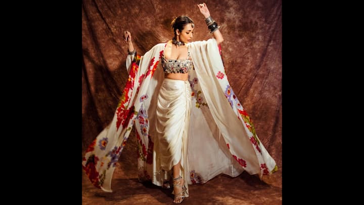 8. विंटेज ग्लैमर वाइब: मलायका अरोड़ा इस आइवरी ड्रेप्ड पहनावे में कालातीत सुंदरता का परिचय देती हैं जो क्लासिक भारतीय सौंदर्यशास्त्र को बोहेमियन परिष्कार के साथ पूरी तरह से मिश्रित करता है। लुक में मिरर-एम्बेलिश्ड क्रॉप्ड चोली के साथ एक रुच्ड साटन स्कर्ट शामिल है। समसामयिक उत्कर्ष को जोड़ते हुए, मलायका ने लाल, बैंगनी और नारंगी रंग के बोल्ड, हाथ से पेंट किए गए पुष्प रूपांकनों से सजी एक बहती हुई हाथीदांत की केप पहनी है। बड़े आकार का केप नाटकीय स्वभाव और हलचल जोड़ता है, जो इस पोशाक को उत्सव की दावतों, मेहंदी समारोहों या गंतव्य शादियों के लिए एक वास्तविक आकर्षण में बदल देता है। (छवि स्रोत: इंस्टाग्राम/@मालाइकारोराऑफिशियल)