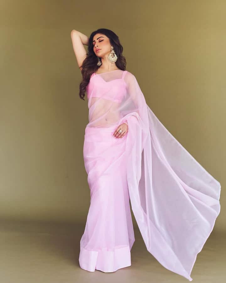In this picture, Mouni is seen in a baby pink colored sheer saree, which looks very delicate and beautiful. She has paired the saree with a matching sleeveless blouse. As accessories, she is wearing big and heavy earrings, which are giving a traditional touch to the look. Her smokey eye makeup and open, wavy hair are completing this graceful and soft look.