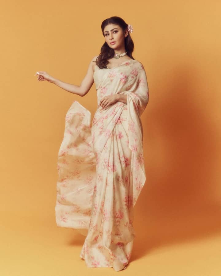 Mouni is in a very soft and feminine look in this look. She is wearing a cream colored floral saree with pink colored printed flowers on it. She has draped the saree in a traditional manner with a sleeveless blouse. As accessories, she wore only a wide pearl choker necklace and a small flower in her hair. This look is graceful, summery and very elegant.