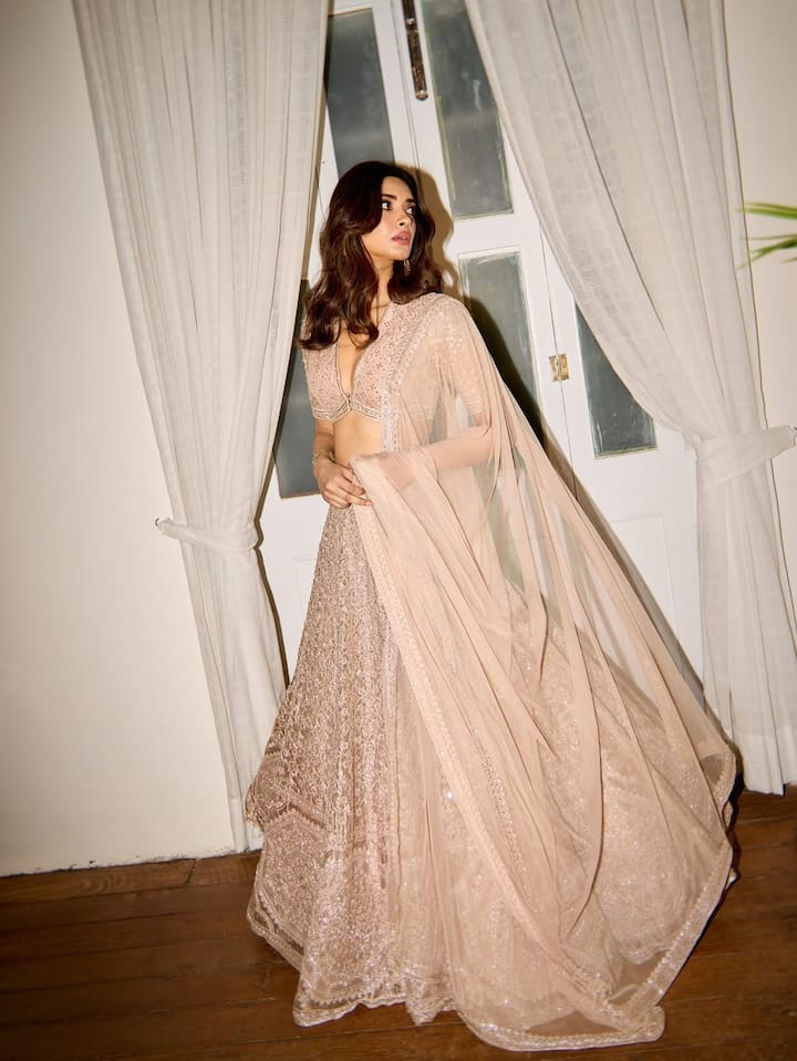 Diana is looking very beautiful and royal in this picture. She is wearing an embroidered lehenga in a light peach-nude shade, paired with a deep neck blouse and a light dupatta. Her open wavy hair and minimal makeup are giving a touch of soft glam to this look. This look looks perfect and elegant for any wedding or festive evening.