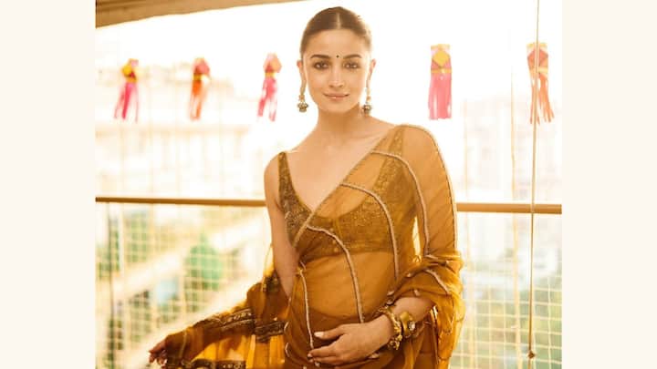 Alia Bhatt glows in this golden mustard organza saree with minimal zari borders, exuding vintage charm. A deep V-neck blouse, sleek bun, and subtle traditional accessories complete her look of quiet sophistication. (Image Source: Instagram/ stylebyami)