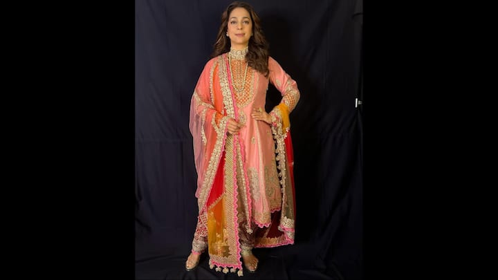 6. Peach And Gold Embroidered Anarkali Ensemble: Juhi Chawla looks ethereal in a stunning peach Anarkali suit adorned with intricate zari and sequin embroidery. The rich craftsmanship on the neckline and sleeves adds a regal touch, while the outfit’s layered flare enhances its traditional grandeur. The vibrant dupatta, featuring shades of red, orange, and yellow with golden lace detailing, adds a burst of colour that beautifully complements the soft peach base. Her look is completed with traditional juttis and a statement choker encrusted with pearls and stones, adding a royal finish. (Image Source: Instagram/@iamjuhichawla)