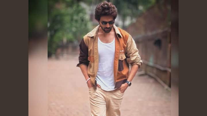 This earthy outfit pairs the white tank with a warm-toned colour-blocked overshirt left casually unbuttoned. Beige trousers and suede boots tie the palette together, giving a rugged yet effortless feel. (Image Source: Instagram/ kartikaaryan)