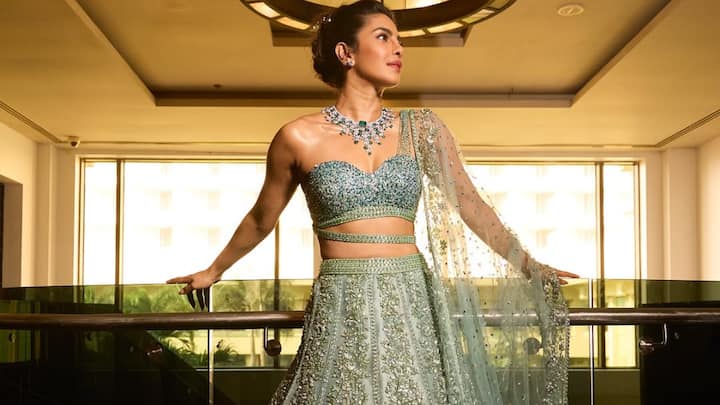 Pastel-Green Celestial Glow: She looks resplendent in this pastel-green lehenga featuring dense floral embroidery that deepens into jewel-toned detailing toward the hem. The hand-worked sweetheart blouse and slim waist belt refine her silhouette, while the sheer sparkling dupatta adds an ethereal finish. Her updo and emerald-diamond necklace amplify the luminous, regal aura. (Image Source: Instagram/ stylebyami)