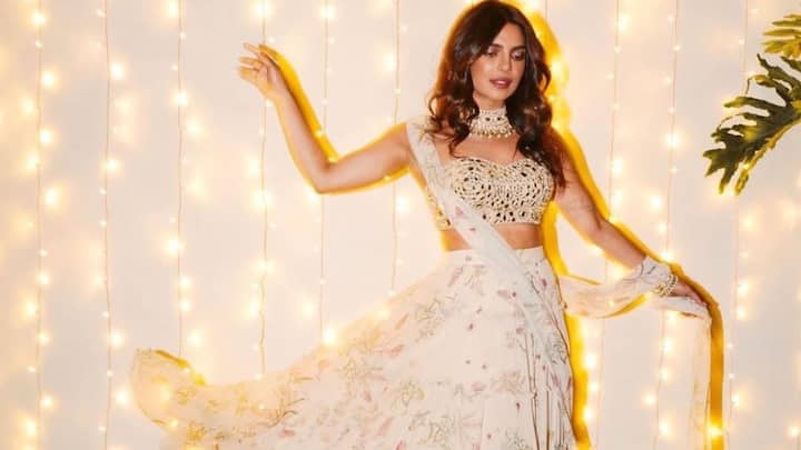 Pastel Dream Lehenga: She looks ethereal in this pastel, multi-layered lehenga that swirls gracefully with its soft floral print. The gold-toned embellished blouse adds a regal contrast, paired with a sheer, easy drape of the dupatta. Her statement choker and soft waves enhance the festive, luminous charm under warm fairy-light glow. (Image Source: Instagram/ stylebyami)