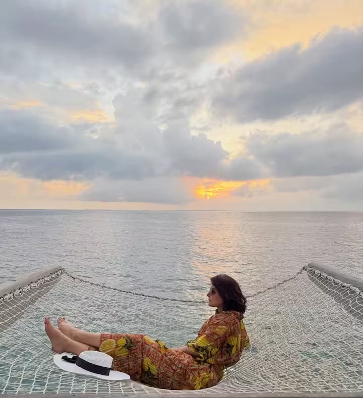 मालदीव की खूबसूरती के बीच हिना खान की यह खूबसूरत तस्वीर उनकी हंसमुख अदाओं को दर्शाती है। समुद्र के किनारे बने एक जाल पर खड़ी हिना ने पीले और लाल रंग से सजी फूलों वाली ड्रेस पहनी है, जो उनके ग्लैमरस और ताजगी भरे लुक को और भी निखारती है। हाथ में टोपी और चेहरे पर धूप का चश्मा लिए उनका लुक छुट्टियों के आनंद और फैशन का बेहतरीन मेल है।