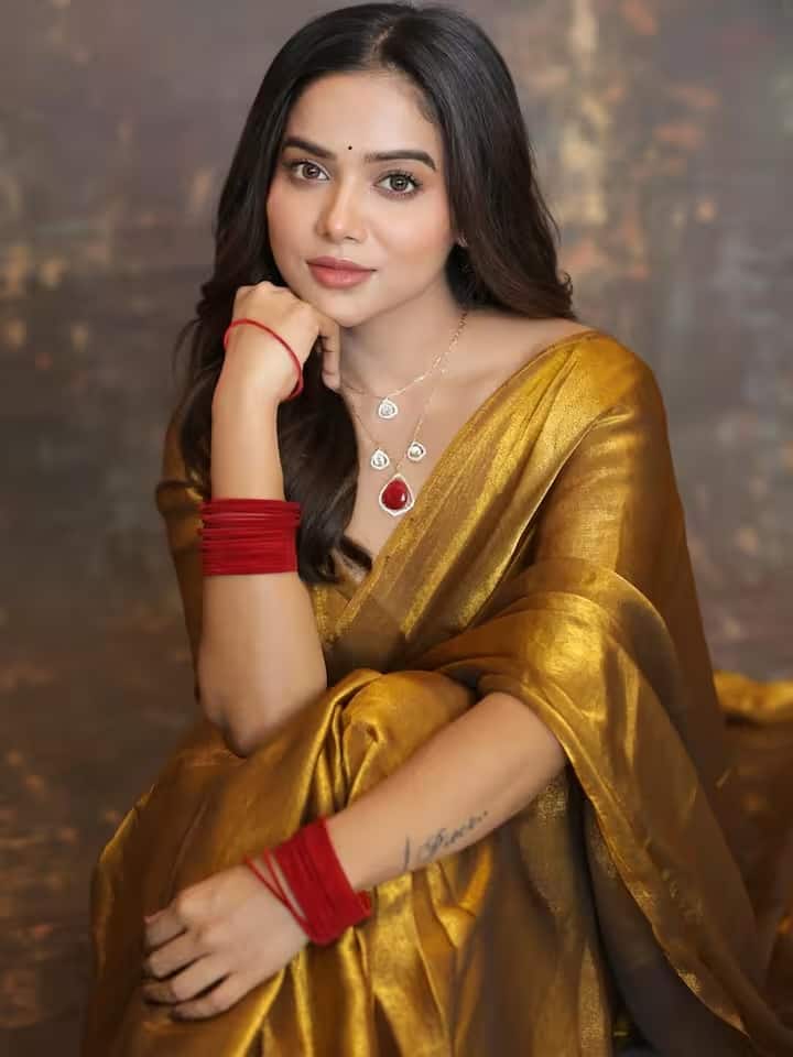 माथे पर लगी छोटी लाल बिंदी और खुले बाल उनके पारंपरिक लुक को खूबसूरती से पूरा करते हैं। लाल और सफेद पत्थरों का हार उनकी शाही साड़ी की शोभा बढ़ाता है।