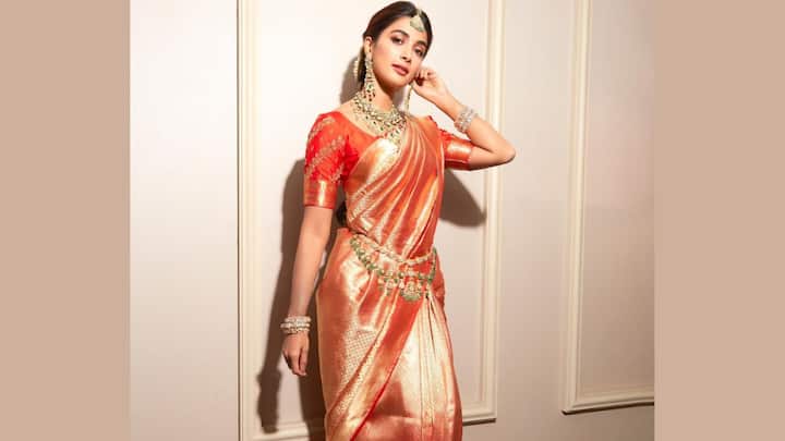Pooja Hegde stuns in this rich copper-orange Kanjivaram silk saree featuring intricate zari patterns that add a luxurious shimmer. Styled with a red blouse, temple jewellery, and a statement kamarbandh, the look radiates perfect bridal-inspired grandeur for wedding festivities. (Image Source: Instagram/ hegdepooja)