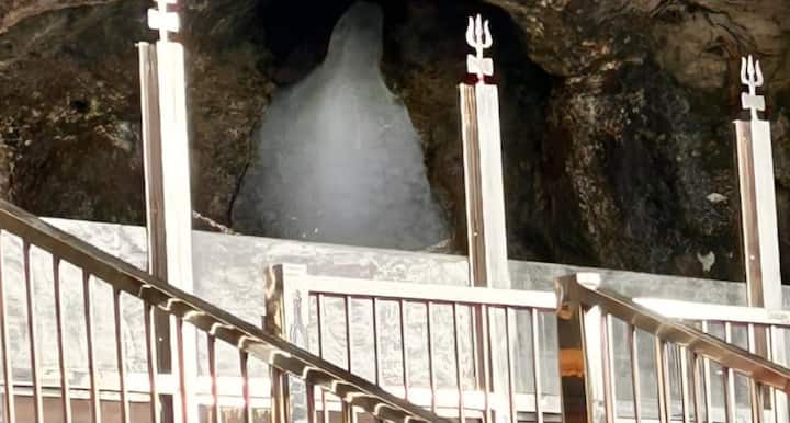 अमरनाथ गुफा के भीतर प्राकृतिक हिमलिंग, जिसका ऊपरी हिस्सा हल्का पिघला हुआ दिखाई दे रहा है. गुफा में श्रद्धालु दूर से दर्शन कर रहे हैं, वातावरण शांत और आध्यात्मिक है. प्राकृतिक रोशनी, यथार्थवादी, सम्मानजनक प्रस्तुति, बिना किसी टेक्स्ट.